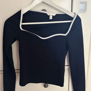 Svart ribbad långärmad topp - Svart ribbad långärmad topp från Bik Bok med vit kant längs den fyrkantiga halsringningen. Snygg croppad passform som ger en modern vibe. Perfekt att styla med jeans eller kjol för en cool look.
