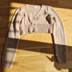 Beige croppad kofta  - Superfin croppad kofta i beige. Stickad med bred ribbad midja, v-ringning och tre stora knappar framtill. Använd någon enstaka gång.