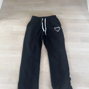 Svarta mjukisbyxor Carsick  - Svarta mjukisbyxor från Carsick i storlek S med vit snörning i midjan och ett broderat hjärta med ansikte på vänster sida. Byxorna har en loose passform och är gjorda i mjukt bomullsmaterial, perfekta för chill dagar. Snygg detalj med det vita broderiet.