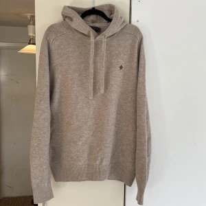 Beige stickad hoodie från Morris - Snygg beige stickad hoodie från Morris med huva och snörning. Tröjan har en broderad logga på bröstet och ribbade muddar vid ärmslut och nederkant. Perfekt för dig som gillar en clean och stilren look. Skriv gärna vid funderingar! 