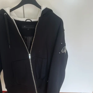 Svart hoodie från Moose Knuckles - Svart hoodie från Moose Knuckles med vit fluffig foder i huvan och quiltad insida. Tröjan har dragkedja framtill, två stora fickor och ett snyggt metallmärke på ärmen. Perfekt för kalla dagar när du vill vara både bekväm och stilren. Samt att Logan är grå och pälsen innuti är vit. Så den skiljer sig lite mer från dem klassiska. 