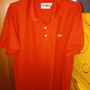 Röd Lacoste pikétröja i bomull - Snygg röd pikétröja från Lacoste med klassisk krage och två vita knappar framtill. Ikoniska gröna krokodilloggan är broderad på bröstet. Tröjan är gjord i mjuk bomull och har korta ärmar, perfekt för en chill och sportig look. 