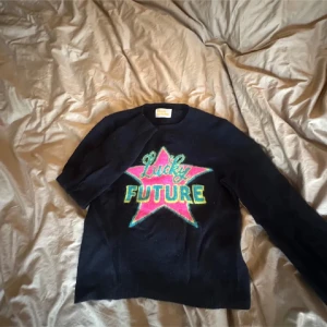Mörkblå kashmirtröja From Future - Superskön mörkblå tröja i 100% kashmir från From Future. Framsidan har en stor stjärna i rosa och grönt med texten 'Lucky Future' i färgglada bokstäver. Tröjan har lång ärm och rund hals, perfekt för dig som gillar statement-plagg och mjuka material.