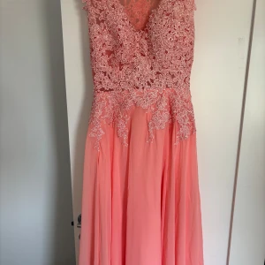 Lång rosa klänning med spets -  En helt oanvänd och jättefin balklänning i ljusrosa/korall med v-ringad spets upptill och blommiga broderier. Kjolen är i flera lager av tunn  polyester.
