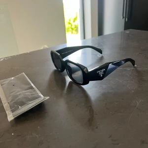 Svarta solglasögon från Prada - Säljer ett par svarta solglasögon från Prada med breda skalmar och rektangulära linser. Glasögonen har en cool, chunky design och logotypen 'PRADA MILANO' i vitt på sidan. Tillverkade i blankt syntetmaterial som ger en lyxig vibe.