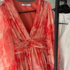 Plisserad blommig klänning från NA-KD - Superfin plisserad klänning från NA-KD i rödrosa och vit med blommigt mönster. Djup v-ringning, långa ärmar och markerad midja. Tunt, flowy material som ger en luftig känsla. Perfekt för dig som vill sticka ut med färg och mönster!