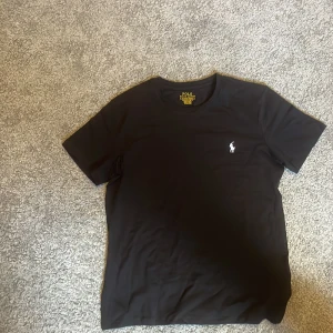 Svart t-shirt från Polo Ralph Lauren - Svart t-shirt från Polo Ralph Lauren med klassisk rund hals och korta ärmar. Slim fit-modell med vit broderad logga på bröstet. Mjuk och skön bomull som sitter snyggt på kroppen. Perfekt för dig som gillar stilrena och enkla plagg.