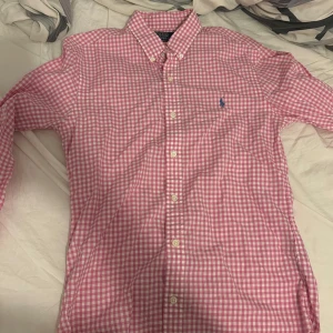 Rosa rutig skjorta från Polo Ralph Lauren - Snygg rosa och vit rutig skjorta från Polo Ralph Lauren med klassisk button-down krage och blå broderad logga på bröstet. Skjortan har långa ärmar och vita knappar hela vägen. Perfekt för en med stil.