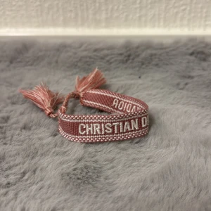 Rosa armband från Christian Dior - Snyggt rosa armband från Christian Dior med broderad logga i vitt och dekorativa tofsar i ändarna. Armbandet har en vävd struktur och är justerbart med knytning. Perfekt accessoar för att lyfta din outfit med lite lyxig vibe. 