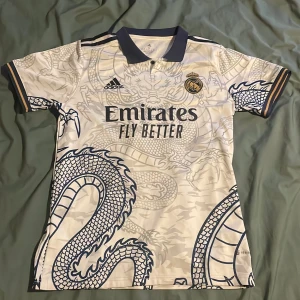 Real Madrid drakmönstrad fotbollströja - Stilren Real Madrid fotbollströja från Adidas i vit med mörkblå detaljer och ett stort drakmönster över hela tröjan. Kortärmad med krage och gulddetaljer på ärmsluten. Materialet är lätt och andas, perfekt för träning, match eller vardagsaktiviteter.