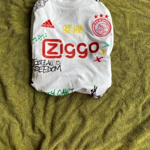 Ajax x Bob Marley Adidas fotbollströja XL - Ajax x Bob Marley special edition fotbollströja från Adidas i vitt med färgglada detaljer och tryck. Tröjan har korta ärmar, reggaeinspirerade ränder på axlarna och flera grafiska prints och citat. Materialet är lätt och andas, perfekt för fotboll eller streetwear.