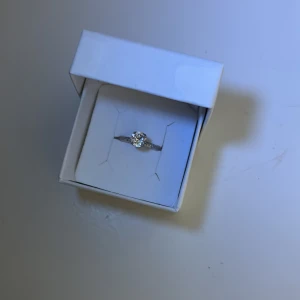 Silverring med kristall från Caroline Svedbom - Snygg silverfärgad ring från Caroline Svedbom med en rund glittrande kristall i mitten. Ringen har en stilren och modern design, perfekt för dig som gillar att sticka ut med eleganta detaljer. Kommer i original ask. Sjustebar, nypris 395kr