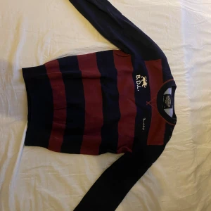 Randig tröja från Bondelid i M - Säljer en snygg långärmad tröja från Bondelid i marinblått och vinrött med breda ränder. Tröjan har broderad logga, Union Jack-flagga på ärmen och förstärkta armbågar. Perfekt för dig som gillar klassisk stil med en sportig touch.