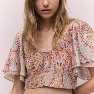 Blommig croppad blus med volangärm - Superfin croppad blus med blommigt mönster i pastellfärger som rosa, lila och beige. Blusen har breda volangärmar, v-ringad hals och öppen rygg med knytband. I helt nyskick aldrig använd men tagit bort prislappen, därav samma pris som nypriset