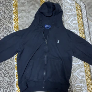 Svart hoodie från Polo Ralph Lauren - Svart hoodie från Polo Ralph Lauren med dragkedja framtill och klassisk vit broderad logga på bröstet. Tröjan har huva, känguruficka och är i bomullsmaterial. Perfekt för en chill och stilren look.