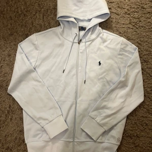 Ljus blå hoodie från Ralph Lauren - Snygg ljus blå hoodie från Polo Ralph Lauren i storlek M. Klassisk design med dragkedja, huva med snörning och broderad logga på bröstet. Tillverkad i mjuk bomull som känns skön mot huden. Perfekt för en chill och stilren look.