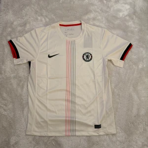 Chelsea bortatröja 25/26 - Chelsea FC vit fotbollströja från Nike med svarta och röda detaljer på ärmarna. Tröjan har tunna vertikala ränder i svart och rött framtill, rund hals och klubbmärke på bröstet. Tillverkad i lätt och ventilerande polyester, perfekt för match eller träning.