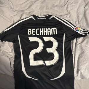 Real Madrid Jersey: Beckham #23  - Säljer en svart Real Madrid Jersey med Beckham 23 på ryggen. Tröjan har vita detaljer, Adidas-logga, klubbmärke och BenQ Siemens som sponsor. Klassisk design med korta ärmar och La Liga-märke på ärmen. Perfekt för dig som älskar fotboll och retrostil.