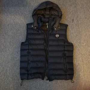 Mörkblå dunväst från Colmar med huva - Snygg mörkblå dunväst från Colmar med avtagbar huva och dragkedja framtill. Västen har två fickor med dragkedja och ett diskret Colmar-märke på bröstet. Västen är storleken L med passar även M nypris 3600 mitt pris 1600 använd endast ett fåtal gånger. Inga skador