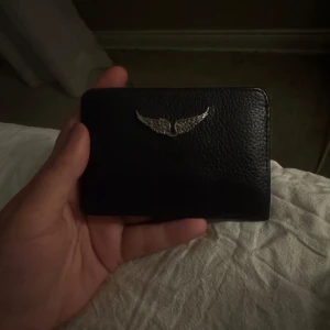 Svart kortfodral Zadig & Voltaire - Stilrent svart kortfodral från Zadig & Voltaire i äkta skinn. Framsidan har en snygg silverfärgad vingdetalj och baksidan har flera kortfack med präglad logga. Perfekt för dig som vill ha ett smidigt och trendigt sätt att bära dina kort.