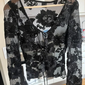 Svart blommig blus - Blommig blus med knyten fram. Jätte fint skick. Använd fåtal gånger. Kommer ej till användning. Genomskinlig men jätte snyggt med bara en bh eller ett linne under. Armarna är lite lösare i slutet vid handen. Är som en storlek xs-s även fast d är M