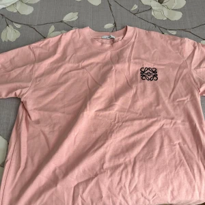 Ljusrosa Loewe t-shirt med logga - Snygg ljusrosa t-shirt från Loewe med svart broderad logga på bröstet. Klassisk rund hals och korta ärmar. Tillverkad i mjuk bomull som känns skön mot huden. Perfekt för dig som gillar stilrena plagg med en twist. Storlek L 