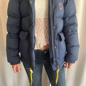 Blå dunjacka från Fjällräven - Säljer en blå, puffig dunjacka från Fjällräven med stor huva och gula snören. Jackan har dragkedja, tryckknappar och två stora fickor framtill. Perfekt för kalla dagar och har en clean, sportig vibe med klassisk Fjällräven-logga på bröstet. Den är i väldigt bra skick och varför jag säljer den är för att jag tänkt köpa en annan färg.