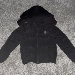 Trappstar jacka - Säljer en svart pufferjacka från Trapstar med huva och dragkedja. Jackan har broderad Trapstar-logga på ryggen och en rund logodetalj på bröstet. Tillverkad i syntetmaterial och har en klassisk pufferlook som är både varm och stilren.