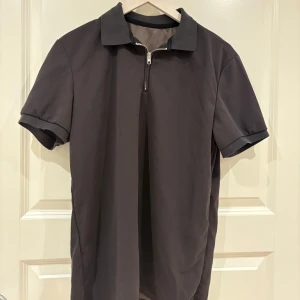 Mörkgrå t-shirt med krage och zip - Snygg mörkgrå t-shirt med korta ärmar, klassisk krage och half zip-dragkedja framtill. Enkel och stilren design som passar till det mesta. Perfekt för dig som gillar en clean look med en twist.