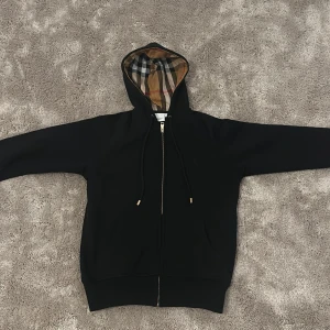 Svart Burberry hoodie  - Svart hoodie från Burberry med klassiskt rutigt foder i huvan och guldfärgade detaljer på snörningen. Tröjan har dragkedja framtill, två fickor och broderad Burberry-logga på bröstet. Perfekt för dig som gillar stilrena och lyxiga streetwear-plagg.