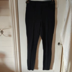 Svarta kostymbyxor  - Svarta kostymbyxor med slimfit passform och pressveck längs framsidan av benen. Byxorna är har hällor för bälte och stängs med knapp och dragkedja. De är i bra skick förutom en lös tråd på framsidan av vänster ben, det vissas i den sista bilden.
