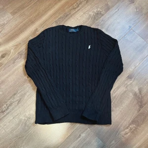 Svart kabelstickad tröja Polo Ralph Lauren - Kabelstickad svart tröja från Polo Ralph Lauren med klassisk rund halsringning och vit broderad logga på bröstet. Långärmad modell i mjuk bomull, perfekt för lager-på-lager och chill stil. Ribbade muddar vid ärmslut och nederkant.