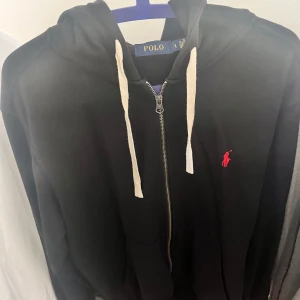 Svart hoodie från Polo Ralph Lauren - Svart hoodie från Polo Ralph Lauren med vit snörning och dragkedja framtill. Klassisk röd logga broderad på bröstet. Tröjan har huva och långa ärmar, samt praktiska fickor framtill. Perfekt för chill dagar och streetwear-stil.