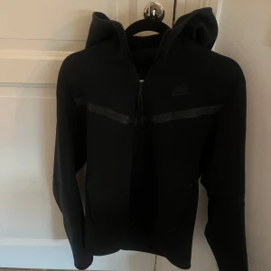 Svart Nike hoodie med dragkedja - Svart hoodie från Nike med hel dragkedja och huva. Snyggt diskret Nike-logga på bröstet och en svart rand över bröstet. Tröjan har långa ärmar och två fickor framtill. Perfekt för chill eller träning.
