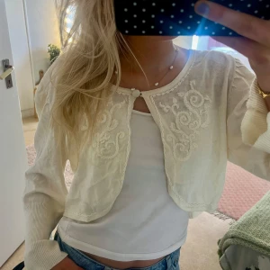 Vit broderad kofta från Soyaconcept - Supersöt vit kofta från Soyaconcept med broderade detaljer framtill och en liten knapp vid halsen. Croppad modell med långärmade puffärmar och ribbade muddar. Perfekt att slänga över ett linne för en chill och feminin look.