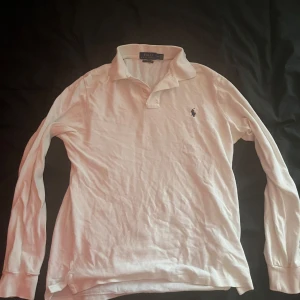 Vit långärmad pikétröja Polo Ralph Lauren - Vit långärmad pikétröja från Polo Ralph Lauren i bomull. Klassisk krage, knäppning vid halsen och den ikoniska broderade loggan på bröstet. Modellen är slim fit och passar perfekt för en clean och stilren look. Pris kan diskuteras.