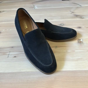 Mörkblå mockaloafers från Geox - Snygga mörkblå loafers från Geox i mjuk mocka med klassisk design och rund tå. Skorna har en stilren ovandel och en ljusbrun sula i syntet. Perfekta för dig som gillar en clean och tidlös look. Använda fåtal gånger