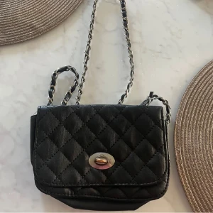 Svart quiltad axelväska med kedja - Snygg svart axelväska med quiltat mönster och silverfärgad kedjerem. Väskan har ett klassiskt lock med vridspänne i metall och är perfekt för att bära det viktigaste. Materialet är mjukt och ger en lyxig känsla.
