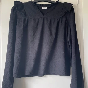 Svart blus med volang från KappAhl - Säljer en svart blus från KappAhl i storlek 170. Blusen har lång ärm, v-ringning och volangdetaljer vid axlar och hals. Väldigt fin och snygg