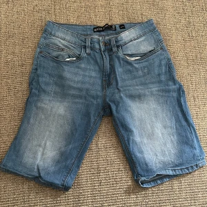 Blå jeansshorts från INDICODE - Snygga blå jeansshorts från INDICODE med klassisk femficksdesign och slitna detaljer. De har normal passform, knapp och dragkedja framtill samt bälteshällor. Perfekta för varma dagar och har en cool, avslappnad vibe.
