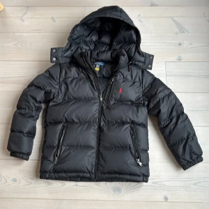 Svart pufferjacka från Polo Ralph Lauren - Säljer en svart pufferjacka från Polo Ralph Lauren med huva och dragkedja framtill. Jackan har två fickor med dragkedja och en liten röd logga på bröstet. Perfekt för kalla dagar och riktigt skön att ha på sig. passar dig som är runt 140-150 cm lång.