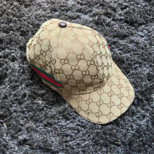 Gucci keps  - Snygg keps från Gucci i beige med klassiskt GG-monogram över hela ytan. Kepsen har gröna och röda ränder på sidan och är tillverkad i bomull. Perfekt accessoar för att lyfta din outfit med en lyxig touch.