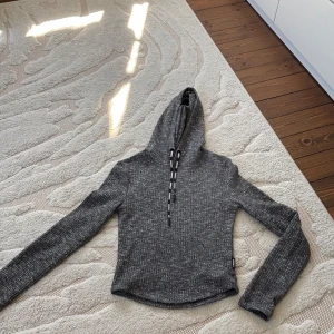 Grå ribbad hoodie från Stronger XS - Sportig gråmelerad hoodie från Stronger i ribbstickat material. Tröjan har långa ärmar, huva med svarta snören där det står 'STRONGER' och en croppad passform. Perfekt för chill eller träning.