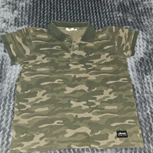 Camo pikétröja från URBNAL - Grön camouflagemönstrad pikétröja från URBNAL med klassisk krage och knappar framtill. T-shirten är kortärmad och har en patch nertill med street style-vibe. Perfekt för dig som gillar en avslappnad och cool look.