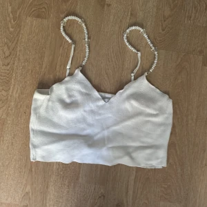 Vit ribbad linnetopp med pärlband - Supersöt vit ribbad linnetopp från New Yorker med unika axelband av pärlor och små fjärilsdetaljer. Croppad modell med v-ringning och stretchigt material som sitter snyggt. Perfekt för dig som vill sticka ut med detaljer och älskar en feminin vibe.
