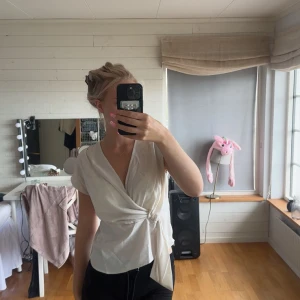 Vit omlottopp med knyt i midjan - Snygg vit omlottopp med korta ärmar och knyt i midjan. Toppen har en v-ringad hals och är tillverkad i ett mjukt, lätt material som ger en stilren och fräsch look. Perfekt för dig som gillar en enkel men trendig stil.