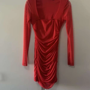 Röd långärmad omlottklänning - Snygg röd kortklänning med lång ärm och djup fyrkantig urringning. Klänningen har ett draperat omlottparti framtill som ger en cool och trendig look. Materialet är lätt och stretchigt, perfekt för dig som vill sticka ut.