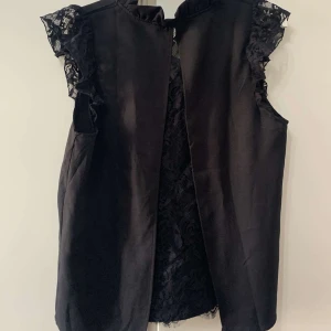 Svart väst med spetsdetaljer - Snygg svart väst med spets på axlarna och längs insidan. Västen är ärmlös och har en öppen framsida, vilket ger en cool och trendig look. Perfekt att slänga över en t-shirt eller skjorta för att lyfta outfiten.