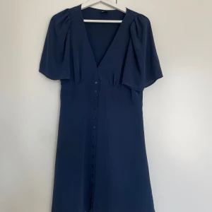 Marinblå klänning från Gina Tricot - Snygg marinblå klänning från Gina Tricot med v-ringning och knappar hela vägen framtill. Modellen har korta, vida ärmar och ett mjukt, flowy tyg som ger en chill vibe. Perfekt för dig som gillar enkel men stilren design.