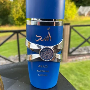 Lattafa Asad Zanzibar parfym - Snygg parfymflaska i matt blått med silverdetaljer och orientaliska mönster. Flaskan har guldtext och ett coolt lejonmotiv framtill. Märket är Lattafa och modellen heter Asad Zanzibar. Perfekt för dig som gillar unika och stilrena dofter. Kan bytas mot någon annan parfymen, eller mot mot parfym som är halva